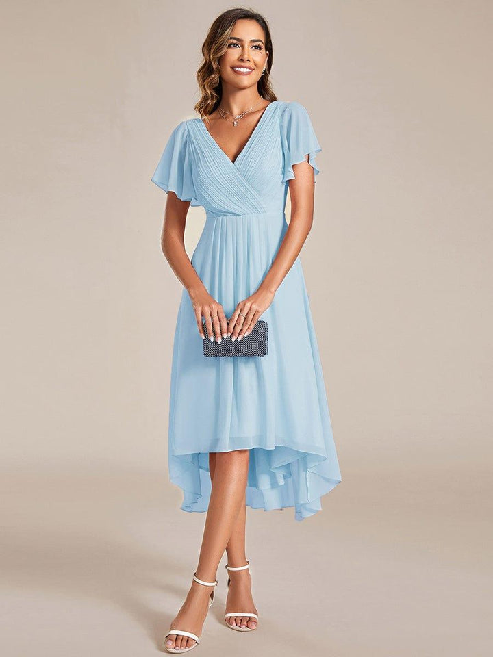 Model holding a small sparkly clutch in the light blue high-low chiffon dress, standing in white heeled sandals. 87