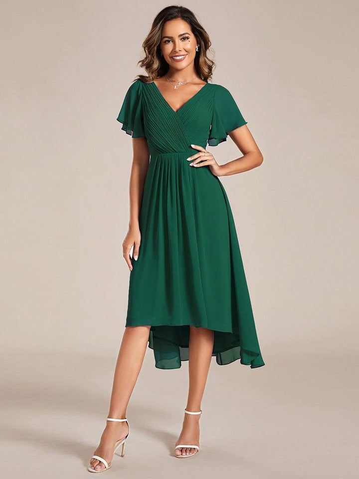 Dark green chiffon dress, front view, with pleated V-neck, flutter sleeves, one hand on hip and white sandals. 10