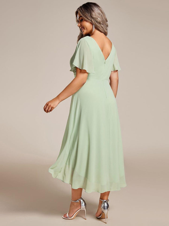 Back view of the sage green chiffon dress on a plus-size model, showing the V-shaped back, zipper and flowing high-low hem. 82