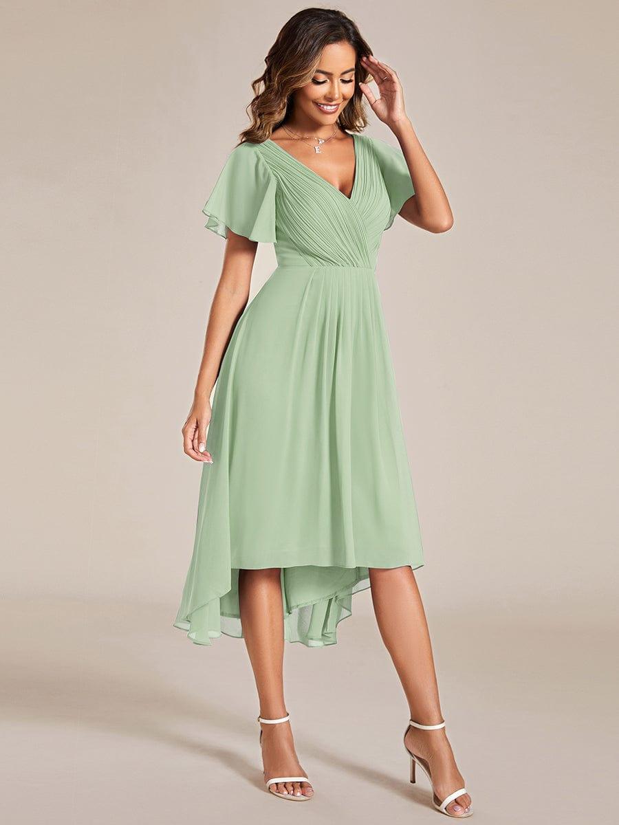 Model in the sage green chiffon dress with one hand raised to her hair, showing the draped high-low skirt and flutter sleeves. 80