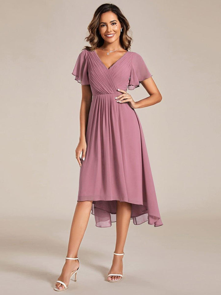 Model in a mauve pink chiffon dress with pleated wrap bodice, flutter sleeves and high-low hem, hand on hip, wearing white heels. 58