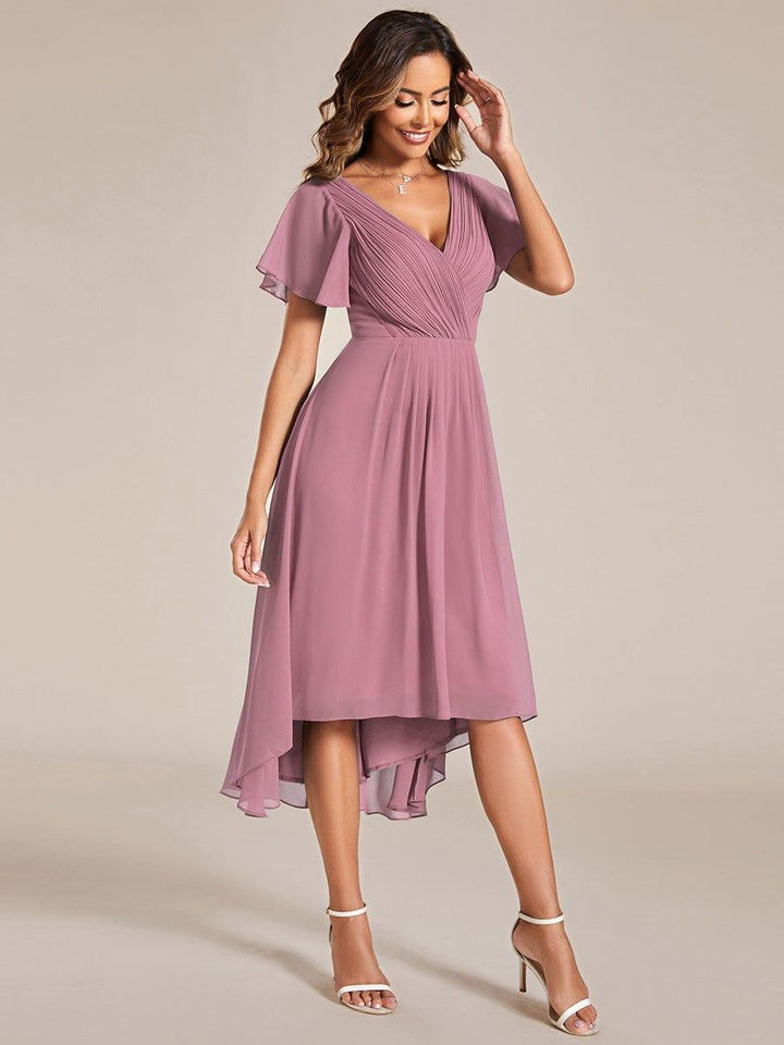 Full-length front view of a model in the mauve pink high-low chiffon dress with white strappy heels, hand raised to her hair. 62