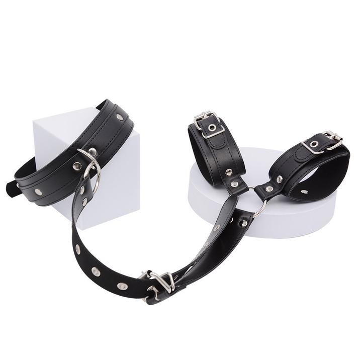 Black collar and wrist cuffs displayed on white blocks and a round platform, joined by a studded strap with silver buckles. 16