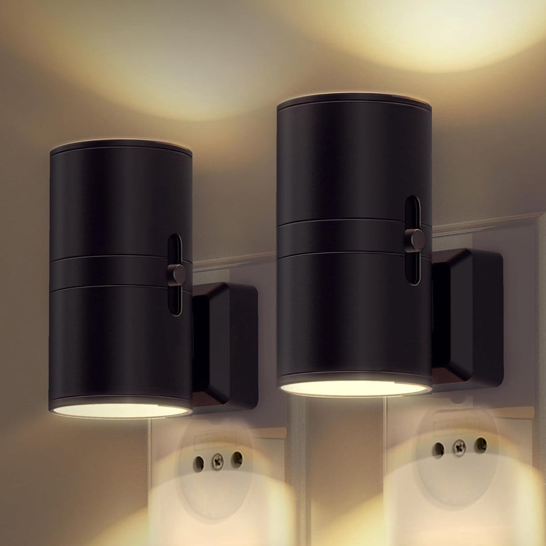 Another view of pair of black cylindrical plug-in lamps with side sliders, glowing warm and lighting the wall above and outlets below.
