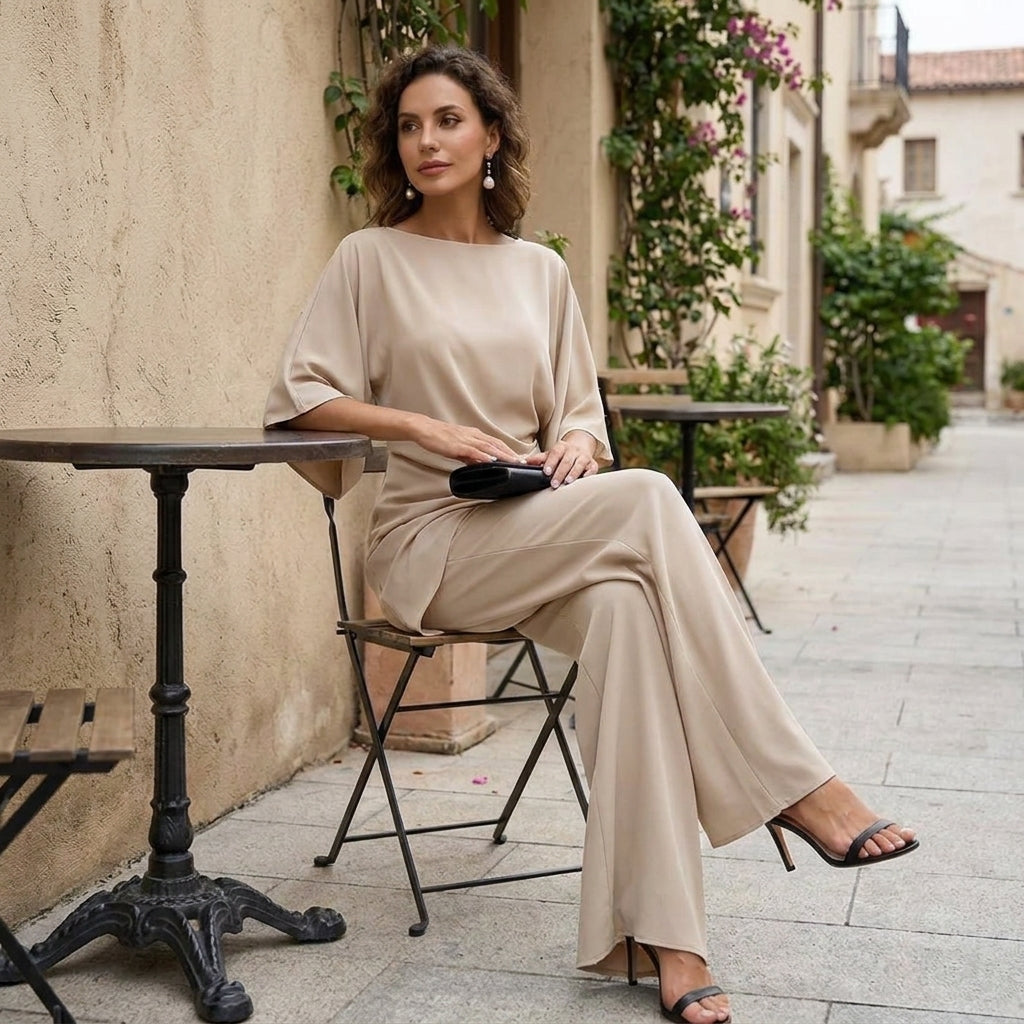 Model in the beige set seated at a small café table on a sunlit patio, legs crossed, wearing black heeled sandals. 12