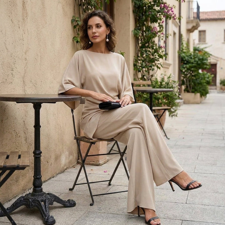 Model in the beige set seated at a small café table on a sunlit patio, legs crossed, wearing black heeled sandals. 12