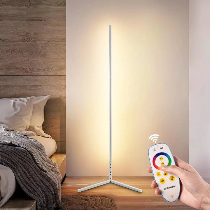 White floor lamp glowing warm beside a bed, with a hand holding the wireless color remote control in the foreground. 9