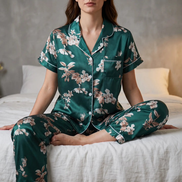 Model sitting cross-legged on a white bed in the green satin short-sleeve pajama set with floral print and white piping. 1
