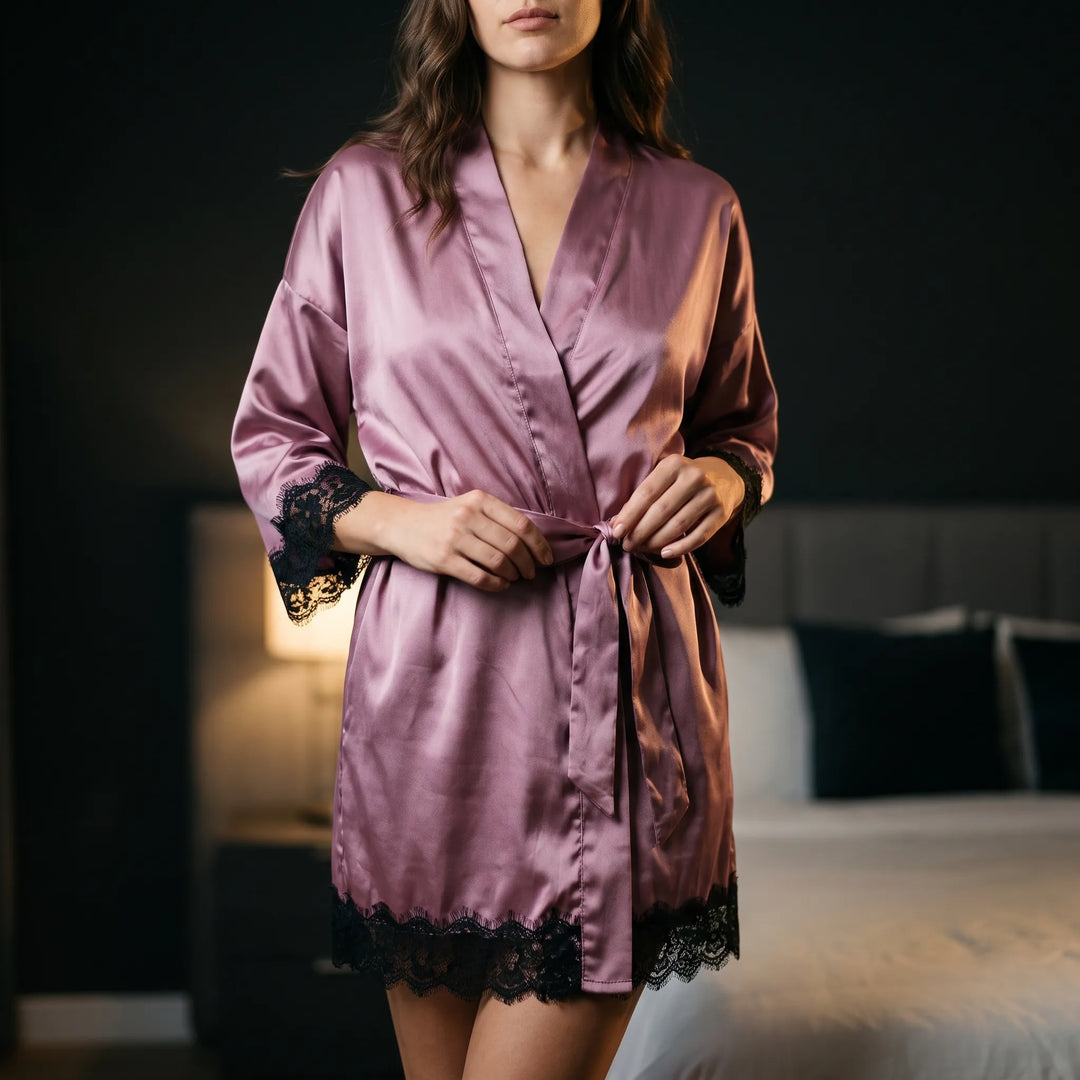 Woman in a mauve satin robe with black lace trim on the sleeves and hem, tying the belt in a bedroom. 1