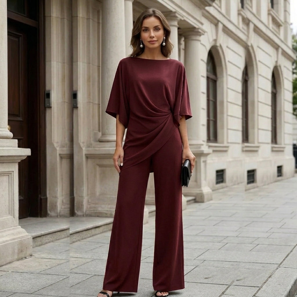 Model in the burgundy set standing in front of a stone building, smiling at the camera, with a black clutch. 11