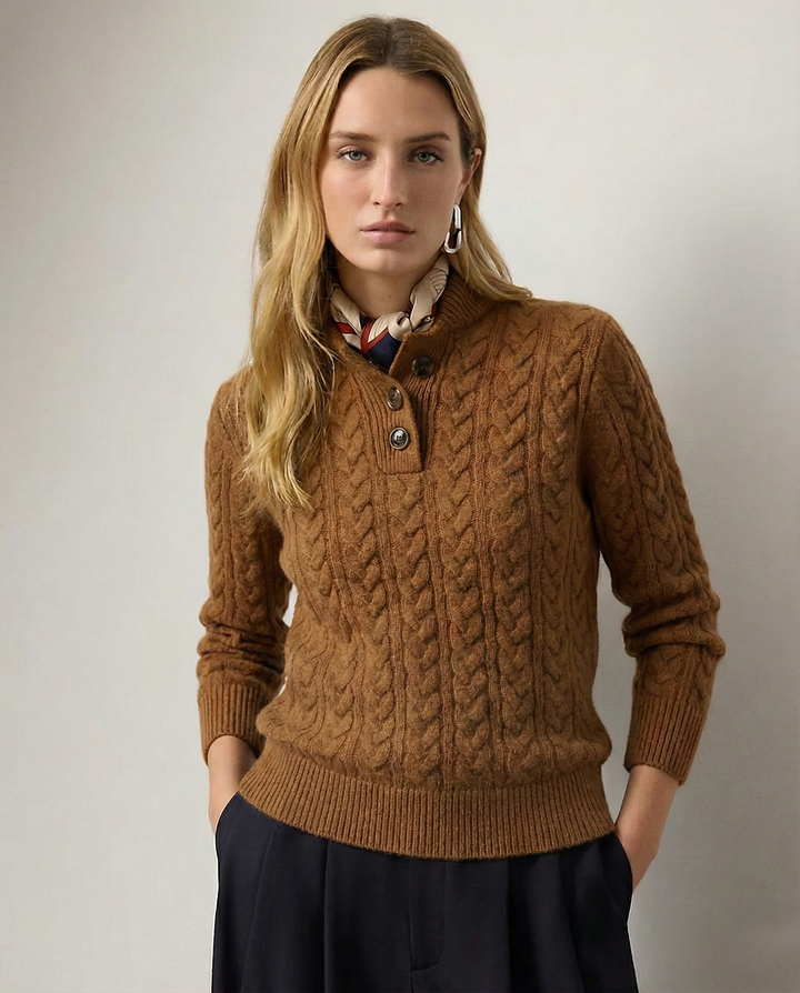 Model facing the camera in the caramel brown cable-knit sweater with three horn buttons, a printed scarf at the collar and dark trousers. 1