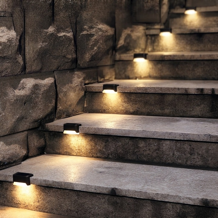 Black solar step lights detail with warm glow lighting rough gray stone steps next to a rugged stone wall.