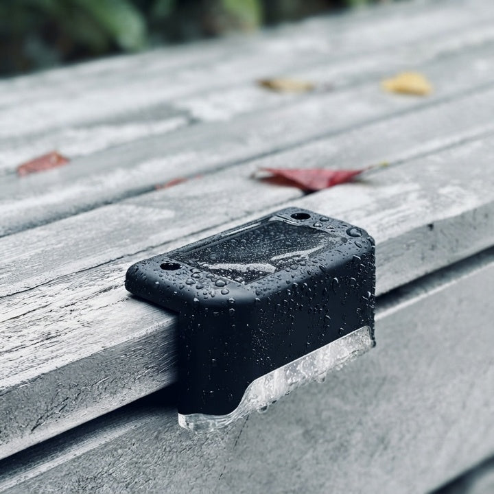 Black solar step light detail on a weathered gray wooden surface covered in water droplets, with fallen leaves nearby.