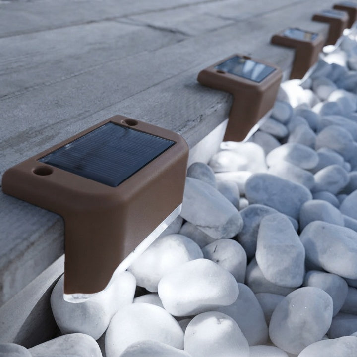 Brown solar step lights detail on a wooden step edge above white decorative pebbles, panels and screw holes visible.