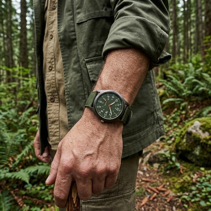 Green military watch with nylon strap, another view worn on a man's wrist in a forest.