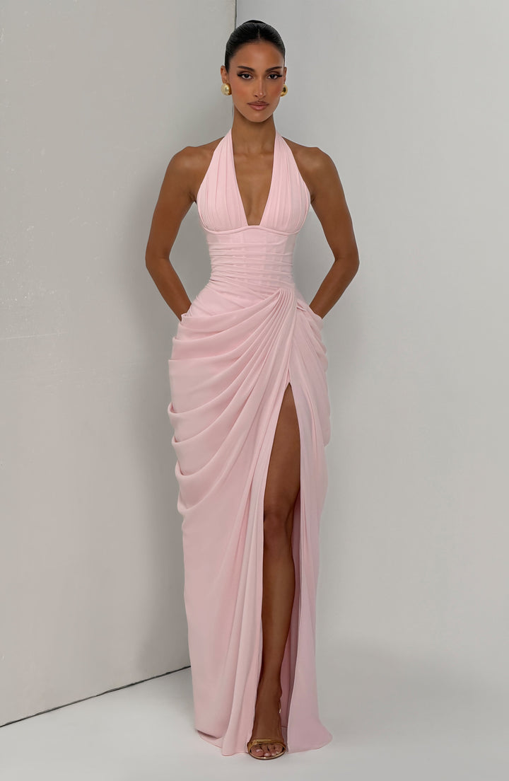 Pale pink halter maxi dress front view against a light gray corner wall with hands in pockets and a high leg slit.