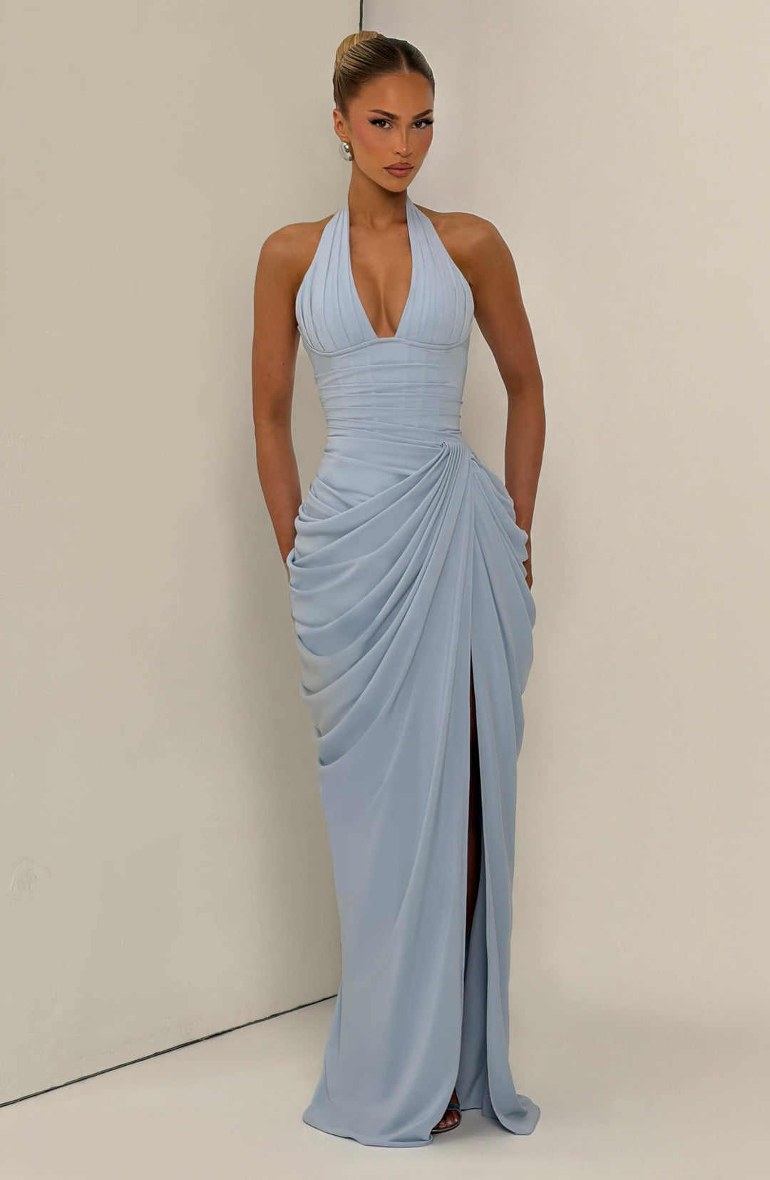 Light blue halter maxi dress front view in a cream corner setting with hands in pockets and the skirt draped to the floor.
