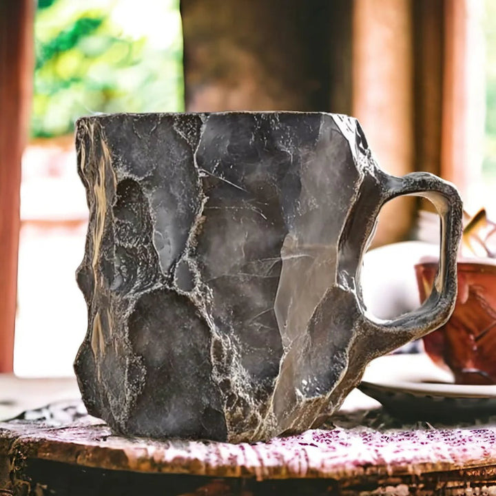 Dark gray mug with faceted, chiseled stone-like surface and a sturdy handle, placed on a patterned cloth near a window. 4