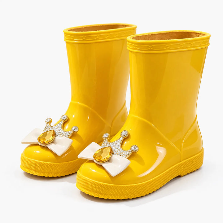 Pair of glossy yellow rain boots at an angle, each toe with a cream bow, jeweled gold crown and amber gem. 10