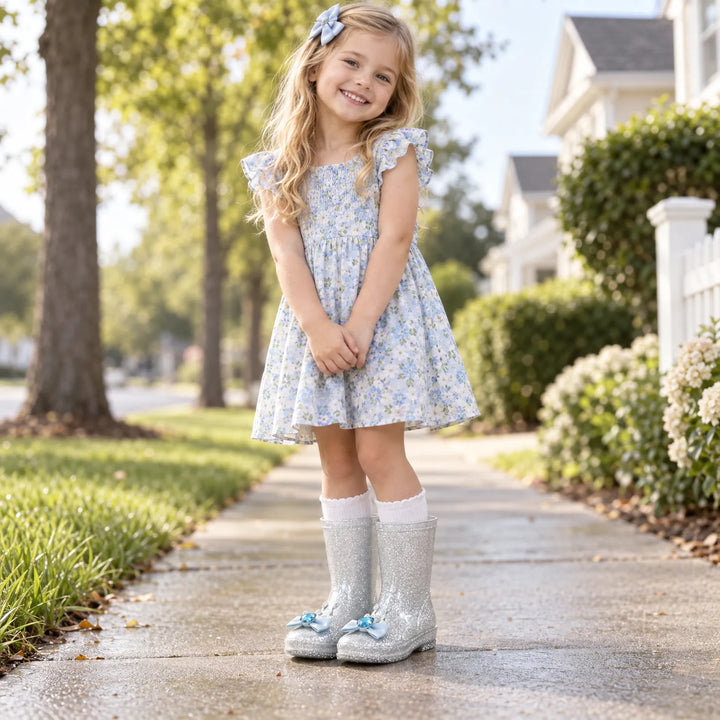 Smiling girl in a floral dress and white socks standing on a wet sidewalk in silver glitter boots with blue gem bows. 3