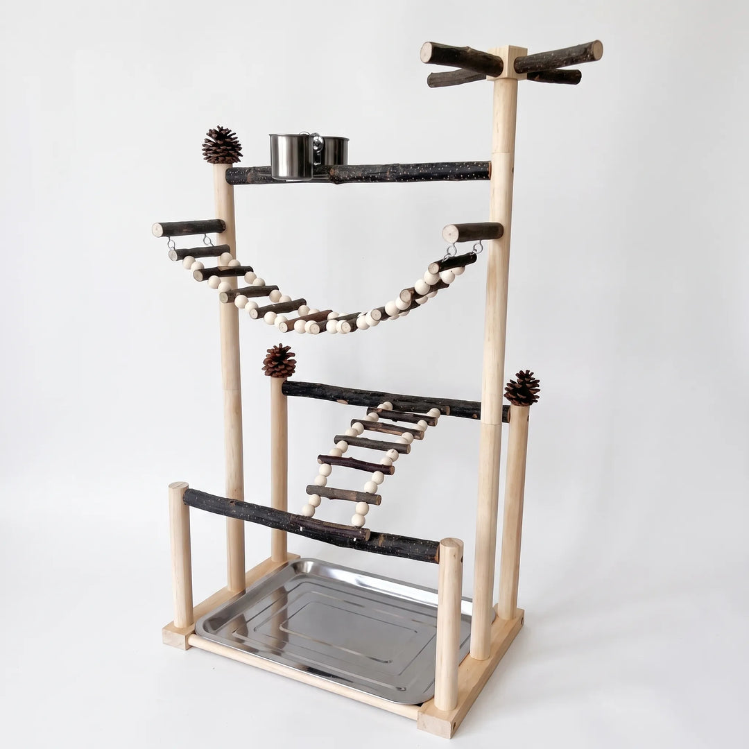 Angled view of a tall wooden bird play stand with natural branch perches, bead-and-twig ladders, pine cones, a steel cup and a steel tray. 1