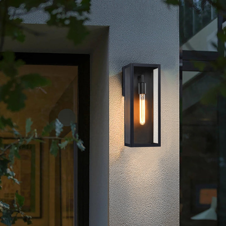 Black outdoor wall lantern with clear glass lit on a gray textured exterior wall next to a doorway.