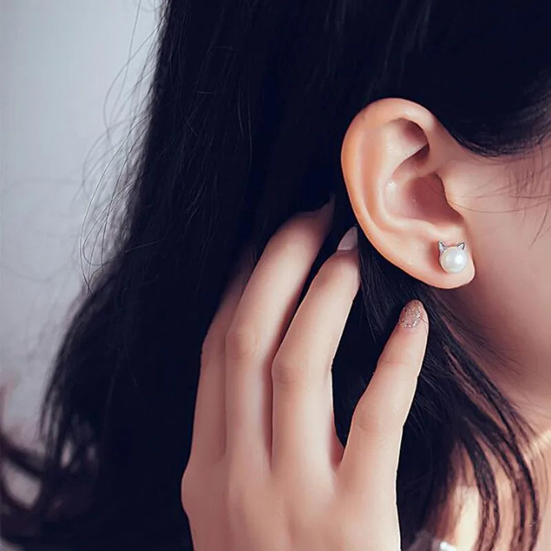 Woman wearing a pearl stud with tiny silver cat ears, with dark hair tucked behind her ear.