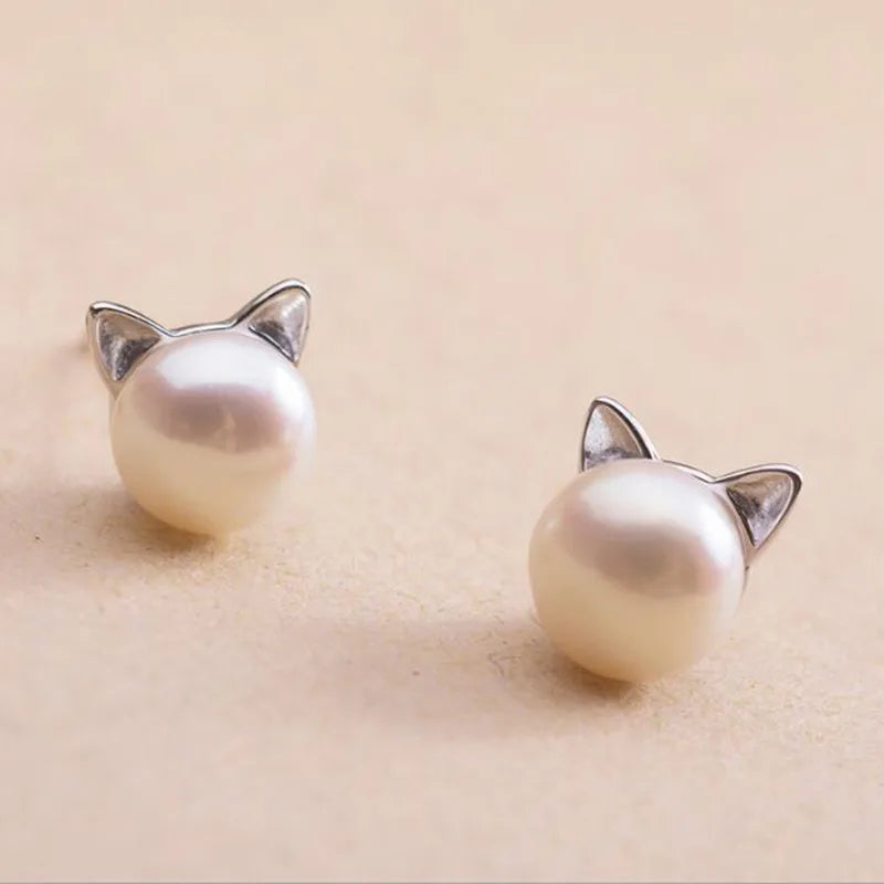 Two pearl earrings with pointed silver cat ears on a soft peach-colored surface.