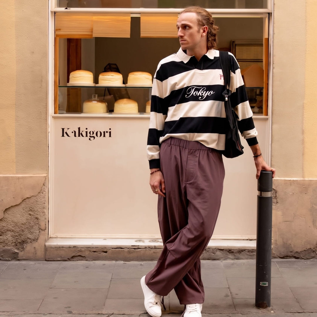 Model in plum-brown wide-leg pants and a striped rugby shirt leaning against a pole outside a bakery window, legs crossed. 3