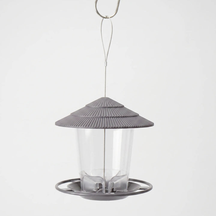 Gray feeder with a ribbed tiered roof, clear seed tube and round tray, hanging from a wire loop and metal hook. 1