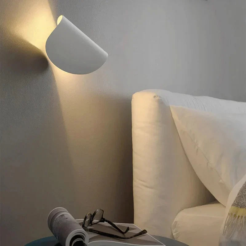 White wall light tilted upward on a bedroom wall beside a cream upholstered headboard, with glasses and a magazine on a nightstand.