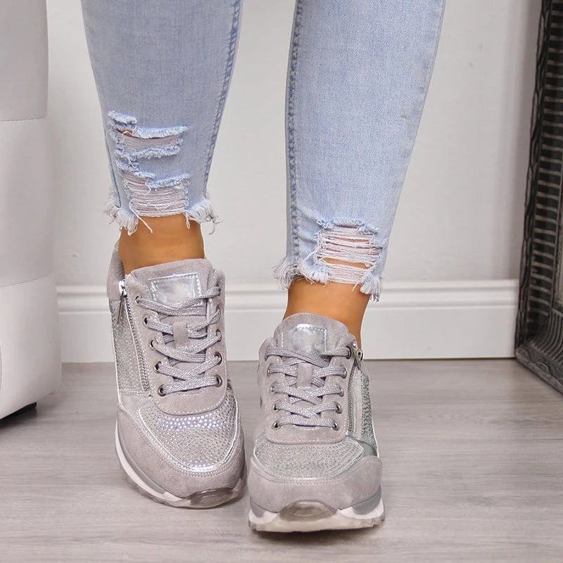 Front view of gray lace-up sneakers with a metallic tongue patch, studded toe panel and outer side zippers.