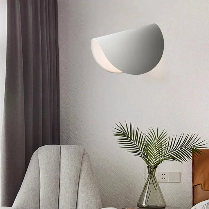 White wall light on a wall above a gray armchair and a glass vase with palm leaves.