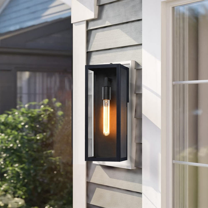 Black outdoor wall lantern with clear glass lit on gray horizontal siding next to a white window frame.