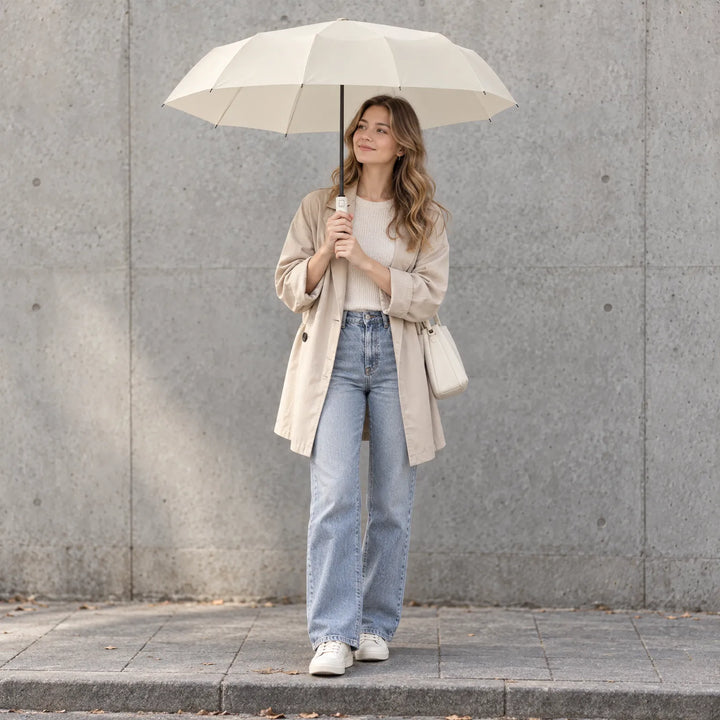 Smiling woman in a beige coat and jeans holds an open cream umbrella while standing before a concrete wall. 1