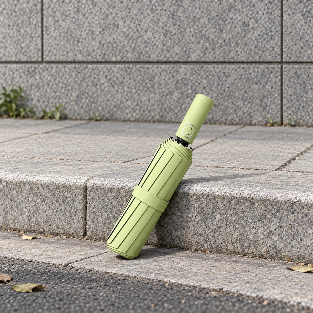 Folded light green umbrella with strap leaning against a granite step on a paved sidewalk, handle tilted upward. 3