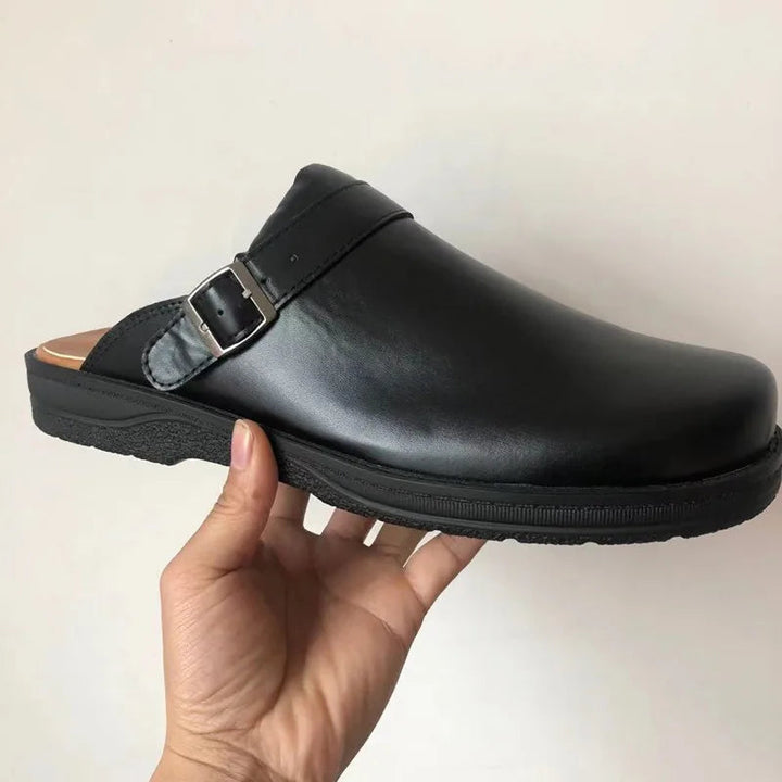 Detail of a single black leather clog in side view with a glossy upper, silver buckle strap and black sole.