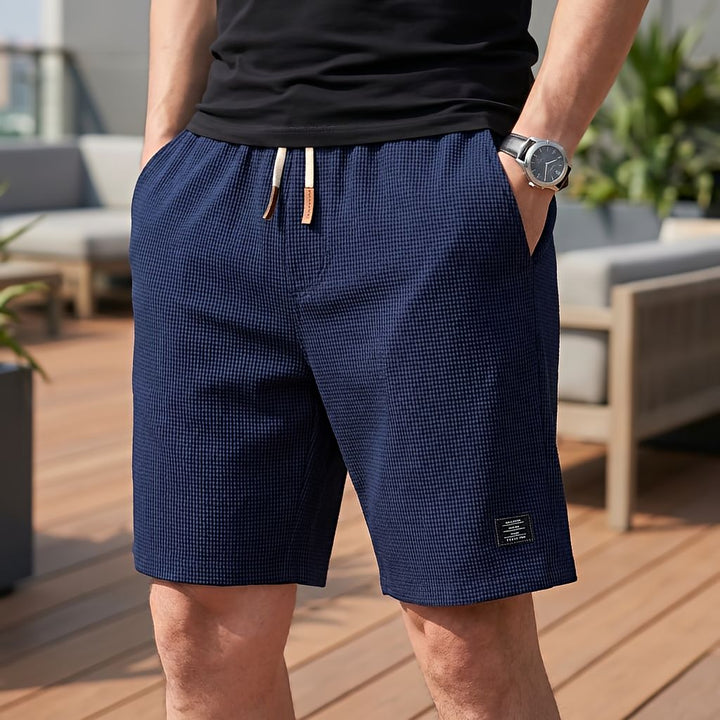 Model wearing navy blue waffle-textured shorts with a white drawstring, hand in pocket, on an outdoor terrace 7