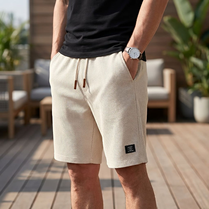 Model wearing beige waffle-textured shorts with a cream drawstring and brown tips, hand in pocket, on a wooden deck 8