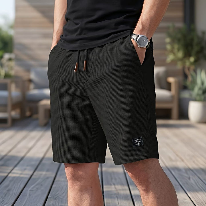 Model wearing black waffle-textured shorts with drawstring and brown tips, one hand in pocket, standing on a wooden deck 3