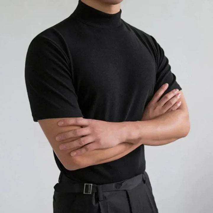 Same black mock-neck tee on a model with crossed arms, shown against a light gray studio wall 2
