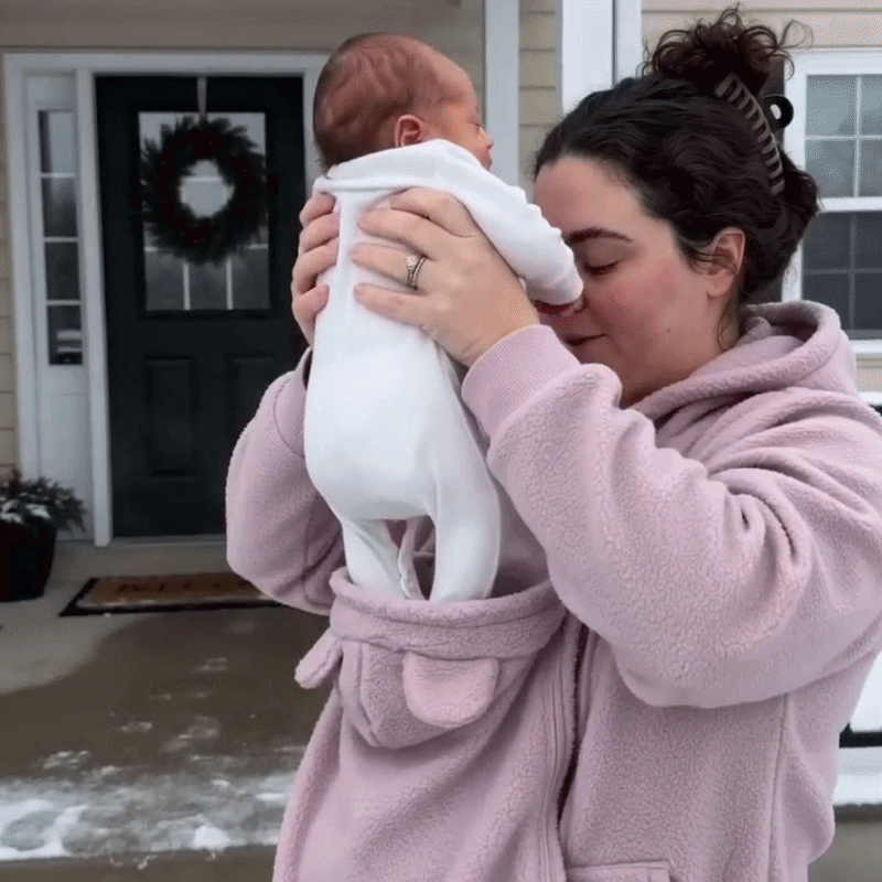 Woman in a pink fleece hoodie lifting a baby in a white outfit out of the front pouch.