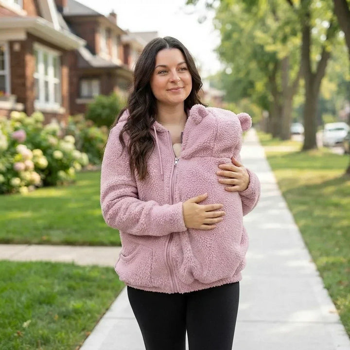 Woman in a pink fleece hoodie with black leggings walking down a sidewalk, holding a baby in the zipped front pouch.