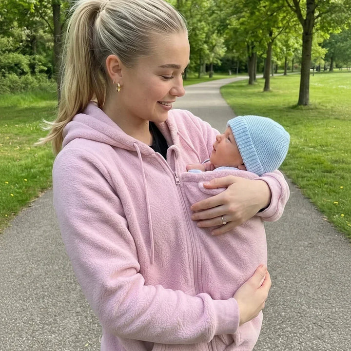 Woman in a pink fleece hoodie holds a baby in a blue knit beanie in the carrier panel.