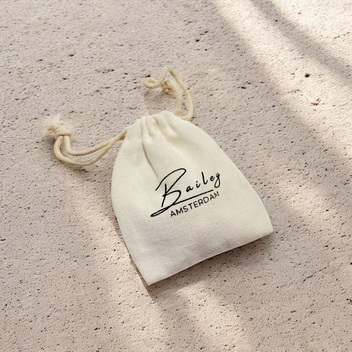 Cream drawstring cotton pouch with black script lettering, lying on a sandy textured surface in dappled sunlight. 8
