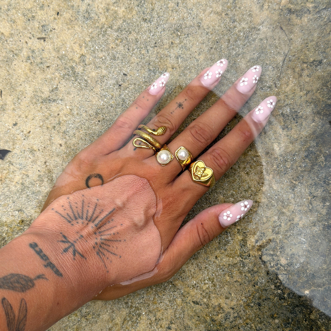 Tattooed hand with flower nails resting in shallow water, wearing the heart ring with a snake ring and pearl rings 4