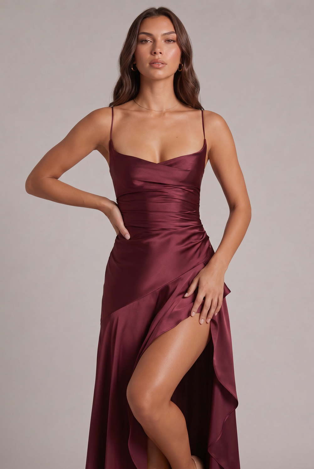 Close-up of the burgundy satin dress bodice, cowl neckline, thin straps and ruffle slit, model with hand on hip 2
