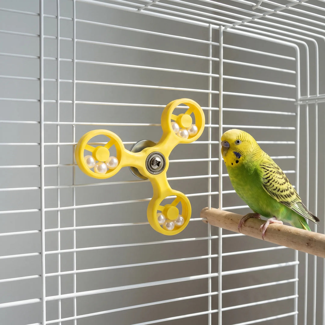 A green budgie on a wooden perch looks at the yellow spinner fixed to the white cage wall. 3