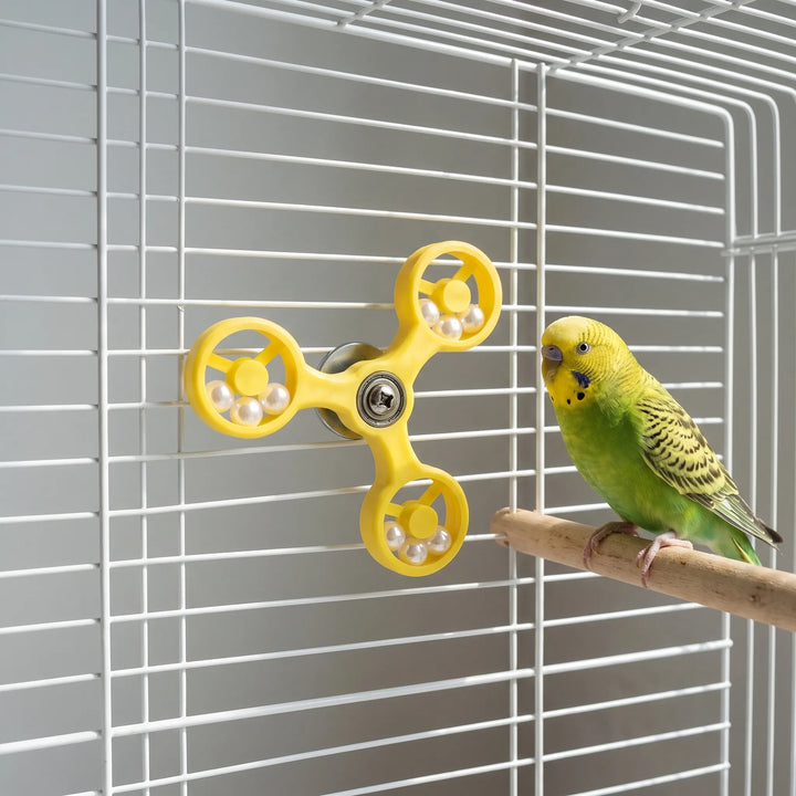 A green budgie on a wooden perch looks at the yellow spinner fixed to the white cage wall. 3