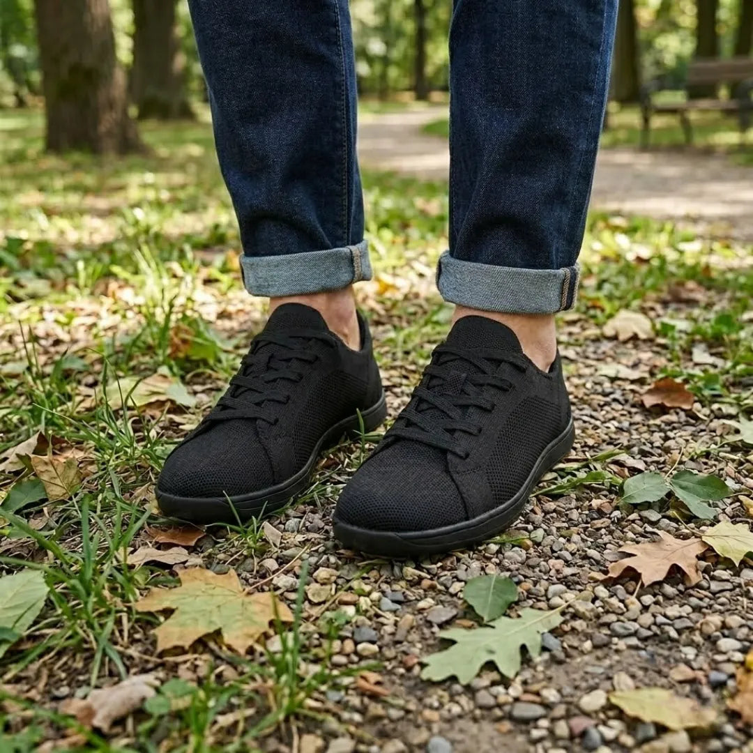 Black knit sneakers worn with rolled dark jeans on a gravel path covered in grass and autumn leaves.